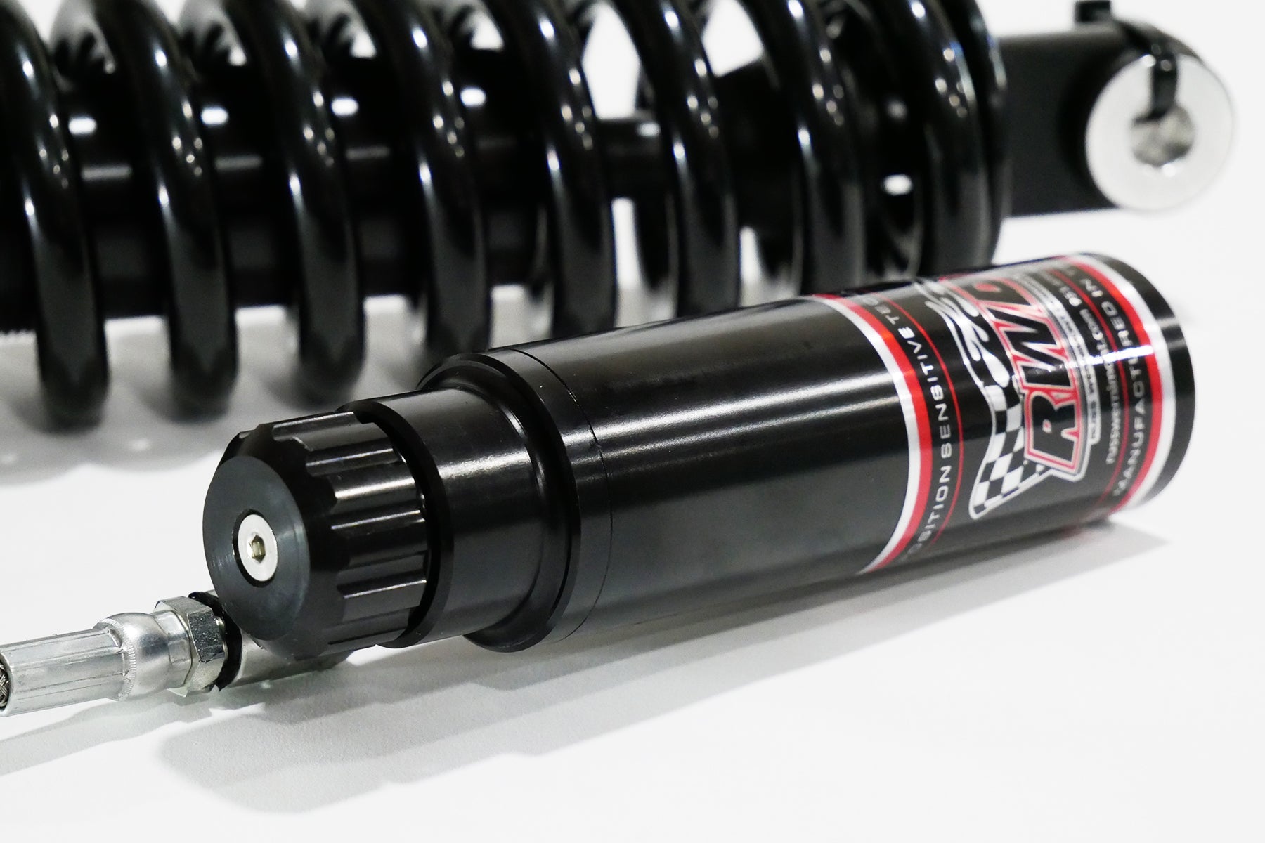 A close-up side shot of an RWD V-Twin RS-2 reservoir, featuring a black body with red and white RWD branding, connected to a silver hose, with a black spring in the background.