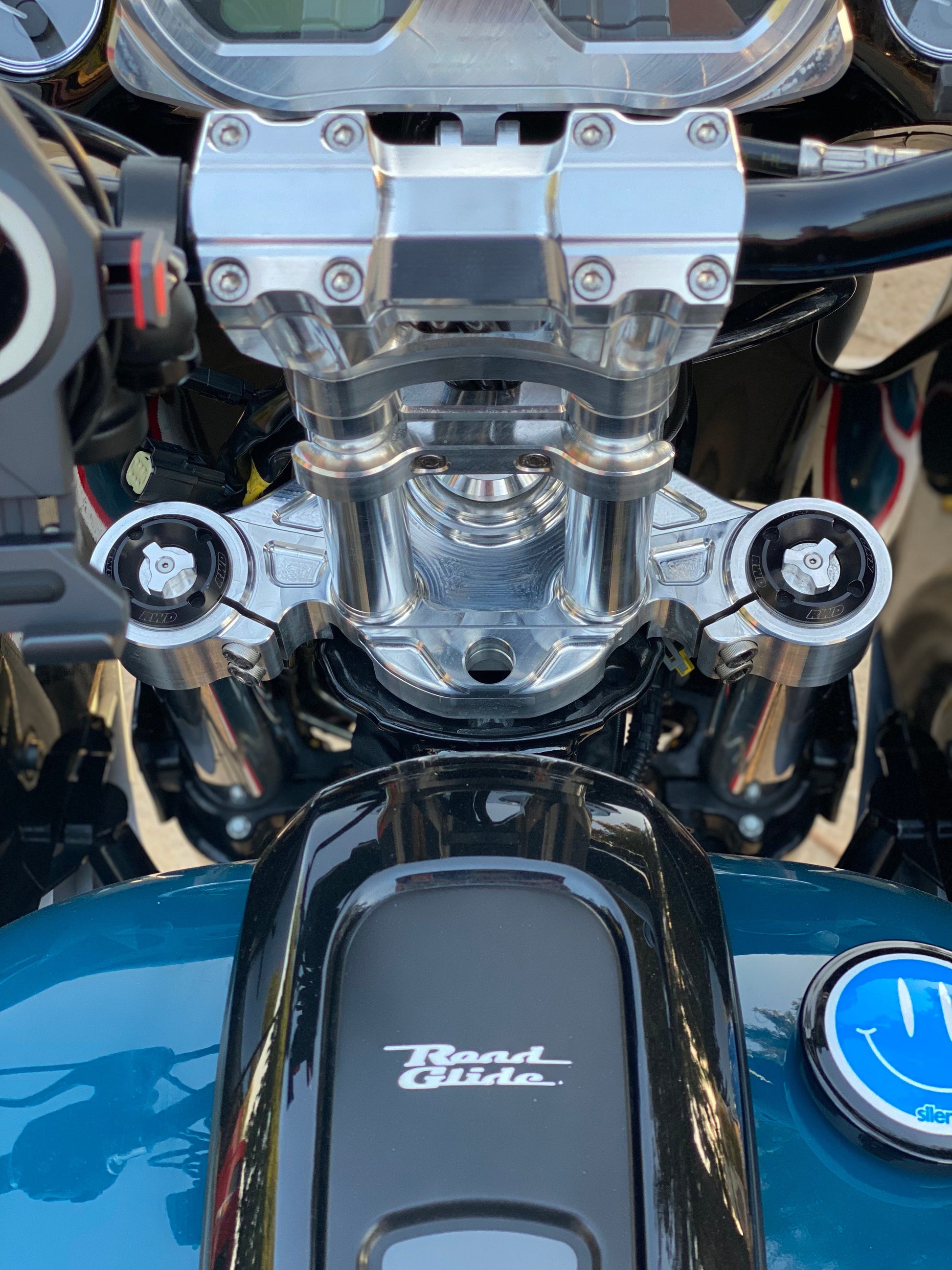 Top-down view of a motorcycle's polished chrome triple tree and black handlebars, featuring RWD V-Twin components, with a black fuel tank displaying the "Road Glide" logo and a blue smiley face cap.